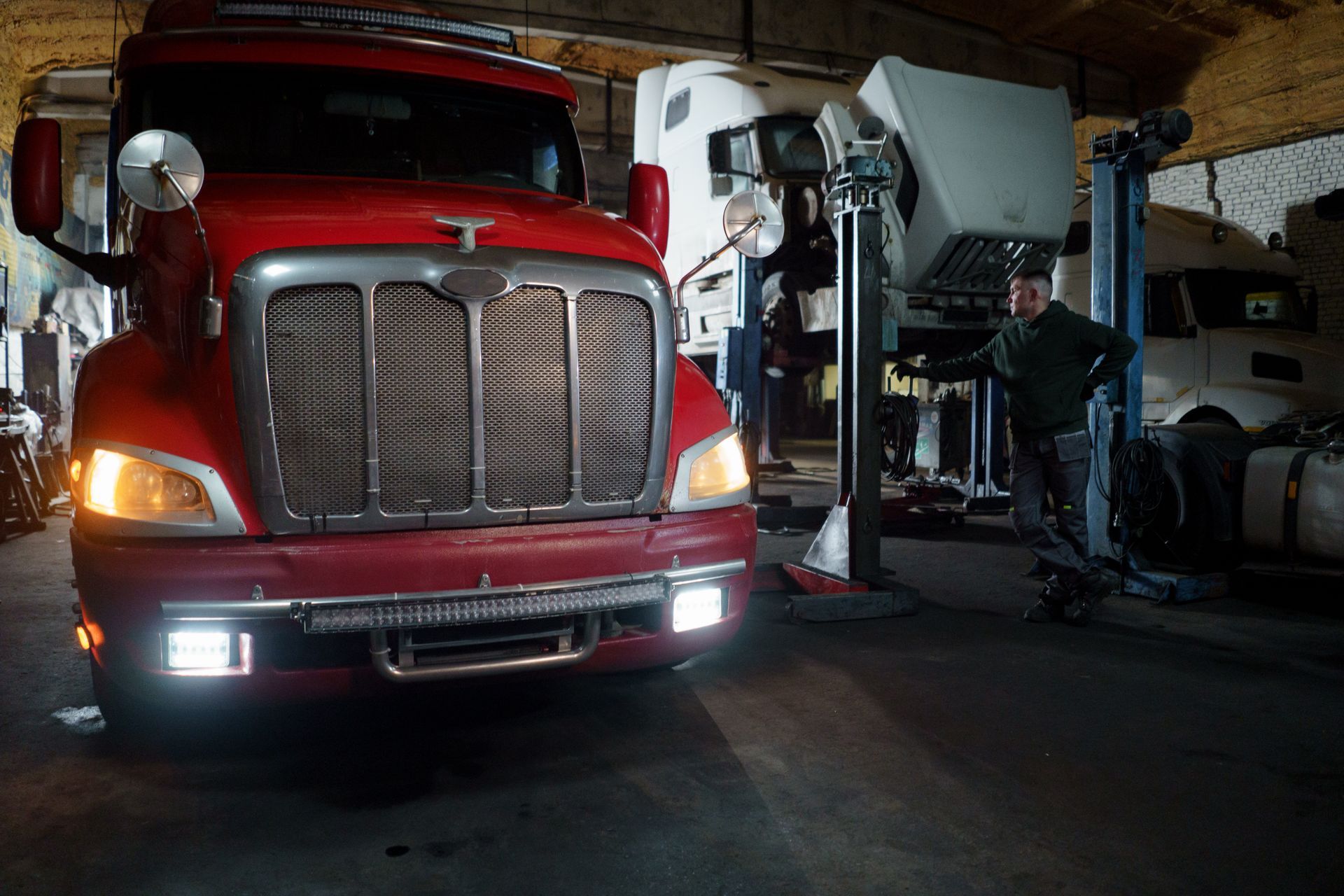 A truck raised on a lift inside a busy repair garage, with a technician working alongside it